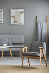 Real photo of armchair standing on carpet and sofa by the wooden coffee table and poster on wall in blurred background