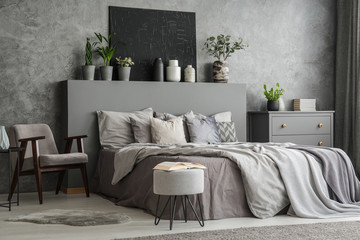 Stylish bedroom interior with bed with blankets and pillows, an armchair, a footstool, a drawer cabinet and a black painting on the wall. Real photo - obrazy, fototapety, plakaty