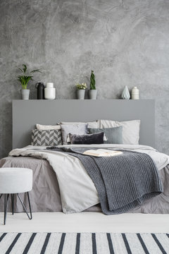 Front view of a comfortable bed with linen, pillows, a blanket and an open book on it in a grey style bedroom interior. Green plants and vases as decorations. Real photo