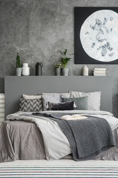 Monochromatic bedroom interior in shades of grey with many cushions and throws on the bed. Vase and plant decorations above the bed. Moon painting on the wall. Real photo