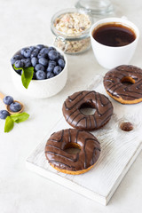 Breakfast concept: donuts with chocolate glaze, a cup of coffee, granola and fresh blueberries with mint on light gray background. Selective focus, copy space 