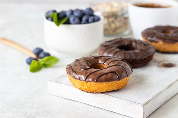Breakfast concept: donuts with chocolate glaze, a cup of coffee, granola and fresh blueberries with mint on light gray background. Selective focus, copy space 