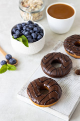 Breakfast concept: donuts with chocolate glaze, a cup of coffee, granola and fresh blueberries with mint on light gray background. Selective focus, copy space 