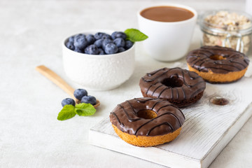 Breakfast concept: donuts with chocolate glaze, a cup of coffee, granola and fresh blueberries with mint on light gray background. Selective focus, copy space 