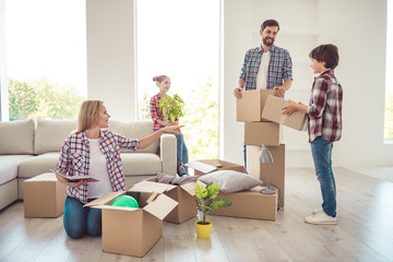 Young happy smiling family four persons unwrapping carton boxes with stuff in light studio living...