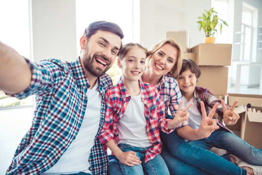 Self portrait of young happy smiling family four persons sitting on floor near carton boxes packages with stuff things in light living room, moving to new flat, showing v-sign gesture