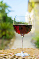 Glass of Red Wine on Rattan Wicker Table in Garden on Villa Mansion. Bright Summer Spring Sunny Day Vegetation Blue Sky. Authentic Lifestyle Image. Relaxation Indulgence Gourmet