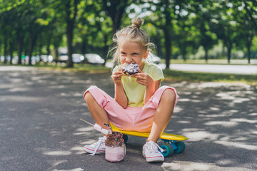 selective focus of little child eating doughnut from dessert while sitting on skateboard at street