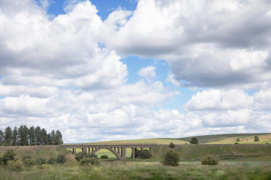 Photograph of a big cloudy sky over rolling green hills and an arched bridge in the Palouse