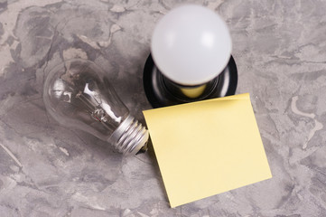 One modern new plastic led electric bulb in black socket and blank yellow curved paper sticker near glass bulb on old worn gray concrete floor