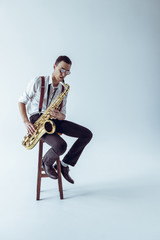 handsome young jazzman sitting on stool and playing saxophone on grey