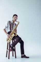 Naklejka premium handsome young performer sitting on stool and playing saxophone on grey