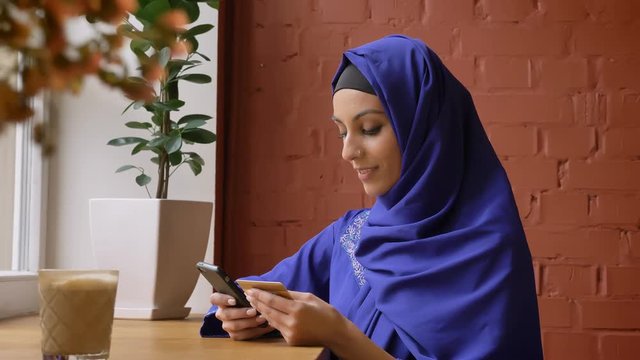 Beautiful young muslim woman in blue hijab rewriting info from card to her phone, sitting in modern cafe