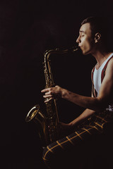 side view of stylish young saxophonist sitting and playing sax on black