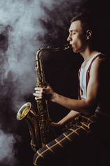 Plakat side view of stylish young jazzman sitting and playing saxophone in smoke on black