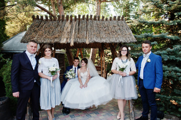 Wedding couple sitting on wooden swings and bridesmaids with groomsmen standing next to them.