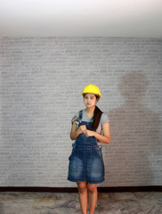 Technician woman ware yellow helmet with grey T-shirt and denim jeans apron dress standing and steel hammer in two hands on grey brick pattern background.