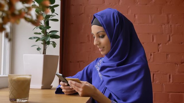 Young charming muslim woman in hijab rewrite info from card to her phone and smiling, sitting in cafe, beautiful female with pierced nose