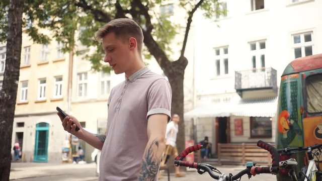 frame from the side happy attractive young man use phone stand with bicycle street mobile love smartphone summer sun tourist adult business hipster caucasian cell office messaging slow motion portrait