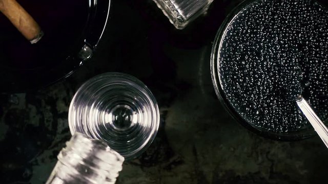 Slow motion pour vodka into a glass next to an ashtray with cigar and black caviar top view