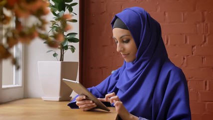 Young muslim woman in blue hijab typing on tablet and sitting in modern cafe, charming lady with pierced nose - Powered by Adobe