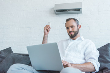 bearded man turning on air conditioner with remote control while using laptop