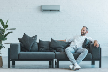 man sitting on grey sofa with remote control, air conditioner on wall