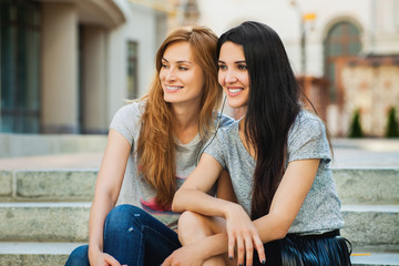 Two young girlfriends sitting on the stairs in the street and listening music. one girl with long brunette hair, another redhead, girls wearing gray shirts. concept of sincere friendship