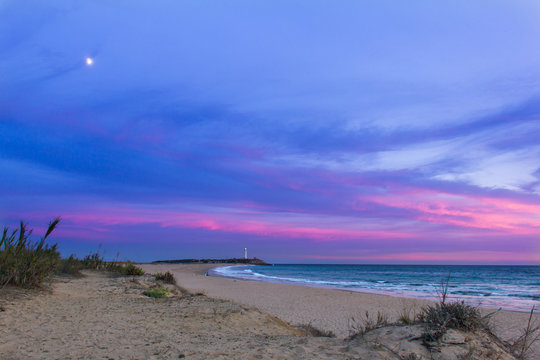 Colorful sunset with moon coming out and Trafalgar lighthouse on the background in empty Zahora beach, Cadiz, South Spain