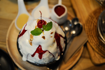 Bingsu milk with sweet red jam on black cup and wooden tray with syrup place on table,A red cherry put on white milk Bingsu,Selective focus