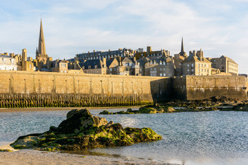 The walled city of Saint-Malo, France, at sunset with the steeple of the cathedral sticking out from the buildings behind the wall and the Fort a la Reine, and a rock in the sea in the foreground.