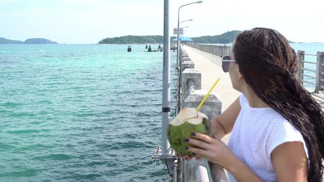 Beautiful girl is drinking coconut on the beach. The girl wears long zizi braids and sunglasses.
