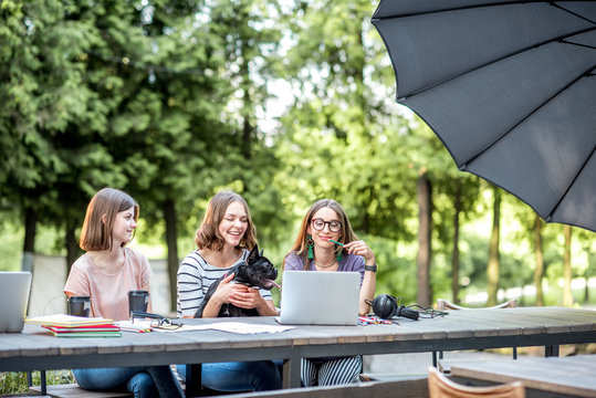 Young Girlfriends Having Fun Together Sitting With Dog At The Outdoor Park Cafe