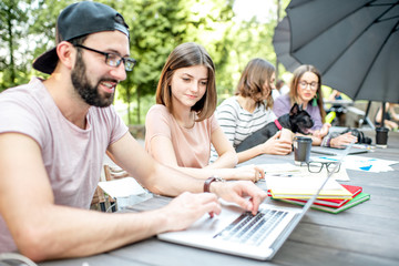 Young people sitting at the big table working or studying with laptops and documents outdoors in the park