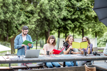 Young friends dressed casually studying with colorful books sitting at the table outdoors in the park
