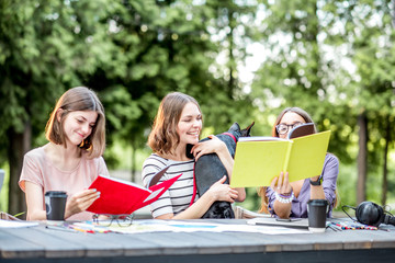 Young friends dressed casually studying with colorful books sitting at the table outdoors in the park