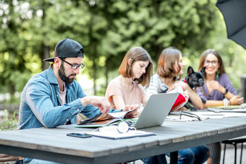 Young friends dressed casually studying with colorful books sitting at the table outdoors in the park