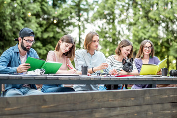 Young friends dressed casually studying with colorful books sitting in a row at the table outdoors