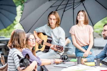 Young friends having fun together playing a guitar sitting at the table outdoors in the park