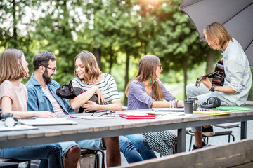 Young friends having fun together playing a guitar sitting at the table outdoors in the park
