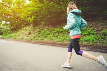 Young fitness blonde woman in headphones running at morning caucasian forest trail in sun light. side view from behind