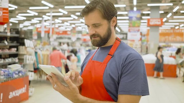 Handsome supermarket clerk using a touch screen tablet in supermarket, he is smiling at camera