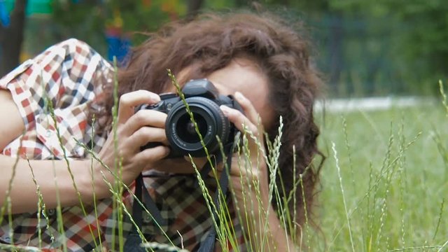 A female photographer is working in the park. A woman with a camera lies on the grass.