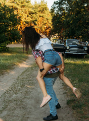 the girl jumped on the guy's back.he holds her legs. check shirt and jeans.in the background is a black car in the forest