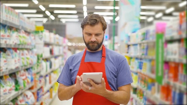 Handsome salesman using digital tablet standing among shelves In supermarket