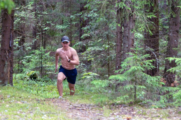 A young man with a trained bare torso, wearing shorts and a cap, runs along the coniferous forest marathon distance