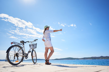 Attractive woman in white closing and sunhat standing at bicycle on stony sidewalk under clear blue...
