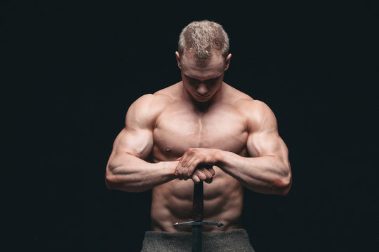 Bodybuilder man posing with a sword isolated on black background. Serious shirtless man demonstrating his mascular body. hands on a sword