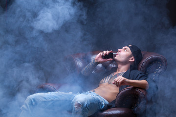Vaper. The man with tattoos sits on a leather sofa smoke an electronic cigarette on the dark...