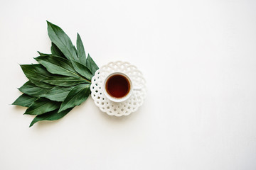 A cup of useful fragrant tasty herbal or black tea on a white surface next to the beautifully laid leaves